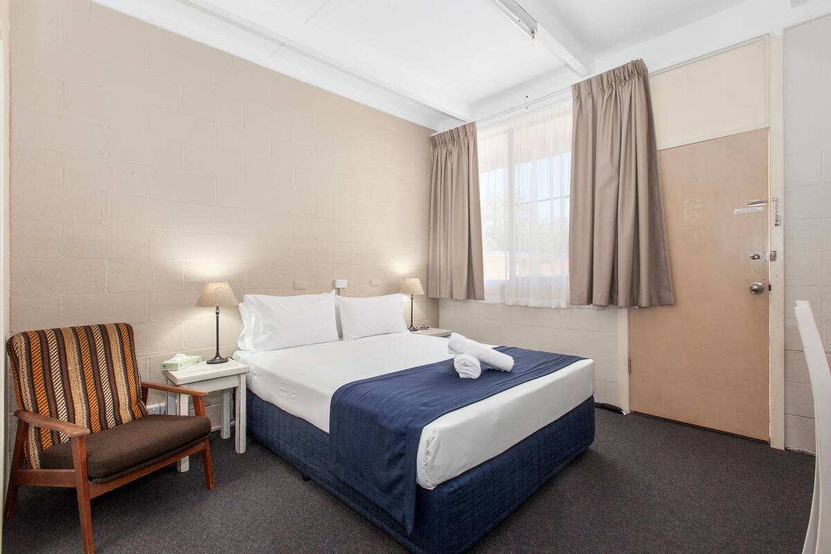 Quarto de hotel com cama, poltrona, mesa de cabeceira, abajur e janela com cortinas.