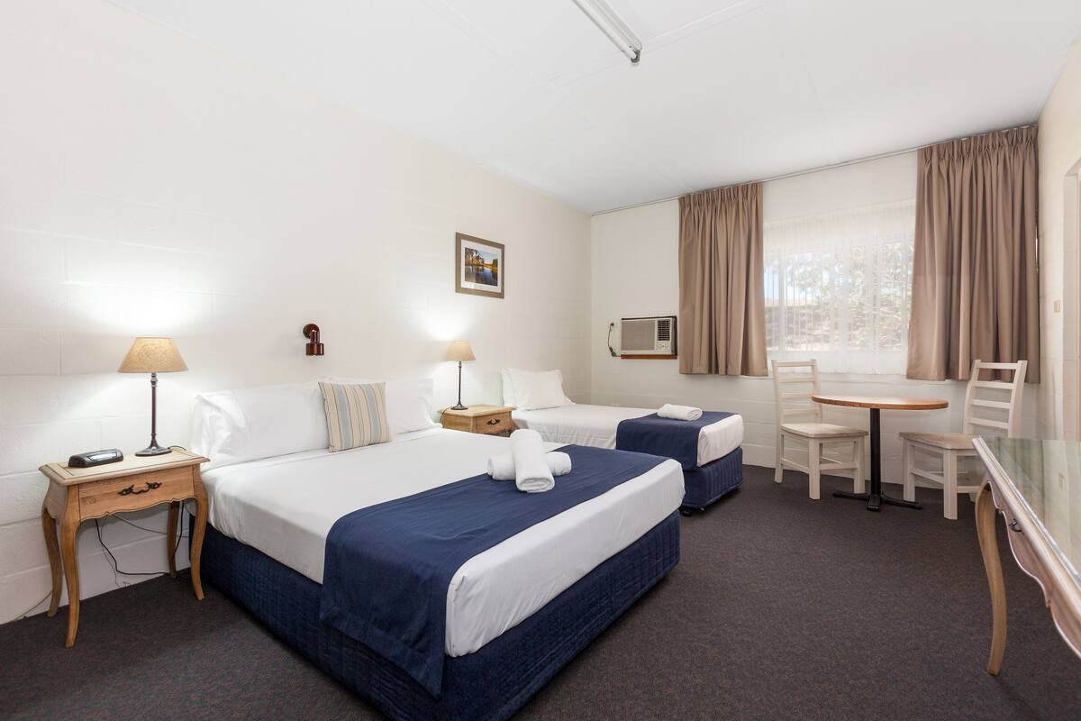Quarto de hotel com duas camas, uma escrivaninha, uma cadeira, um abajur, um quadro na parede e uma janela com cortinas.
