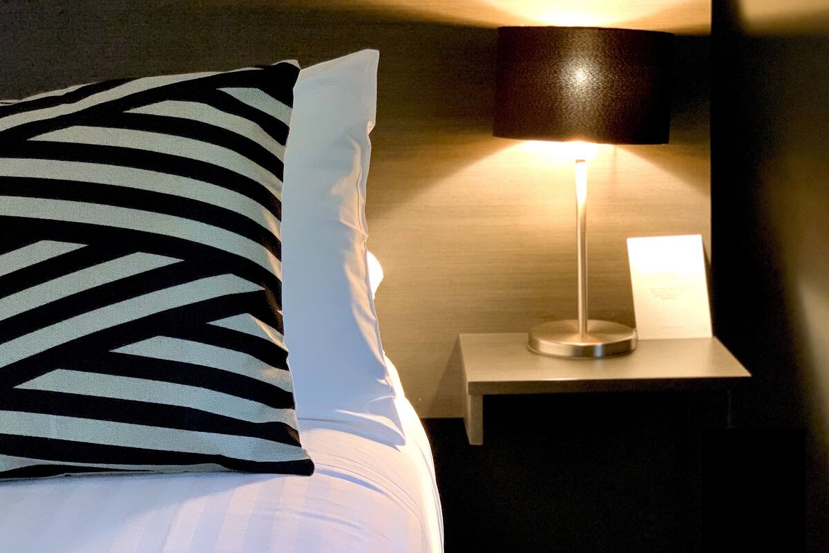 Ein ordentlich bezogenes Bett mit einem gestreiften Kopfkissen und einem weißen Kissen, eine Nachttischlampe und ein kleiner Tisch mit einer darauf stehenden Lampe.