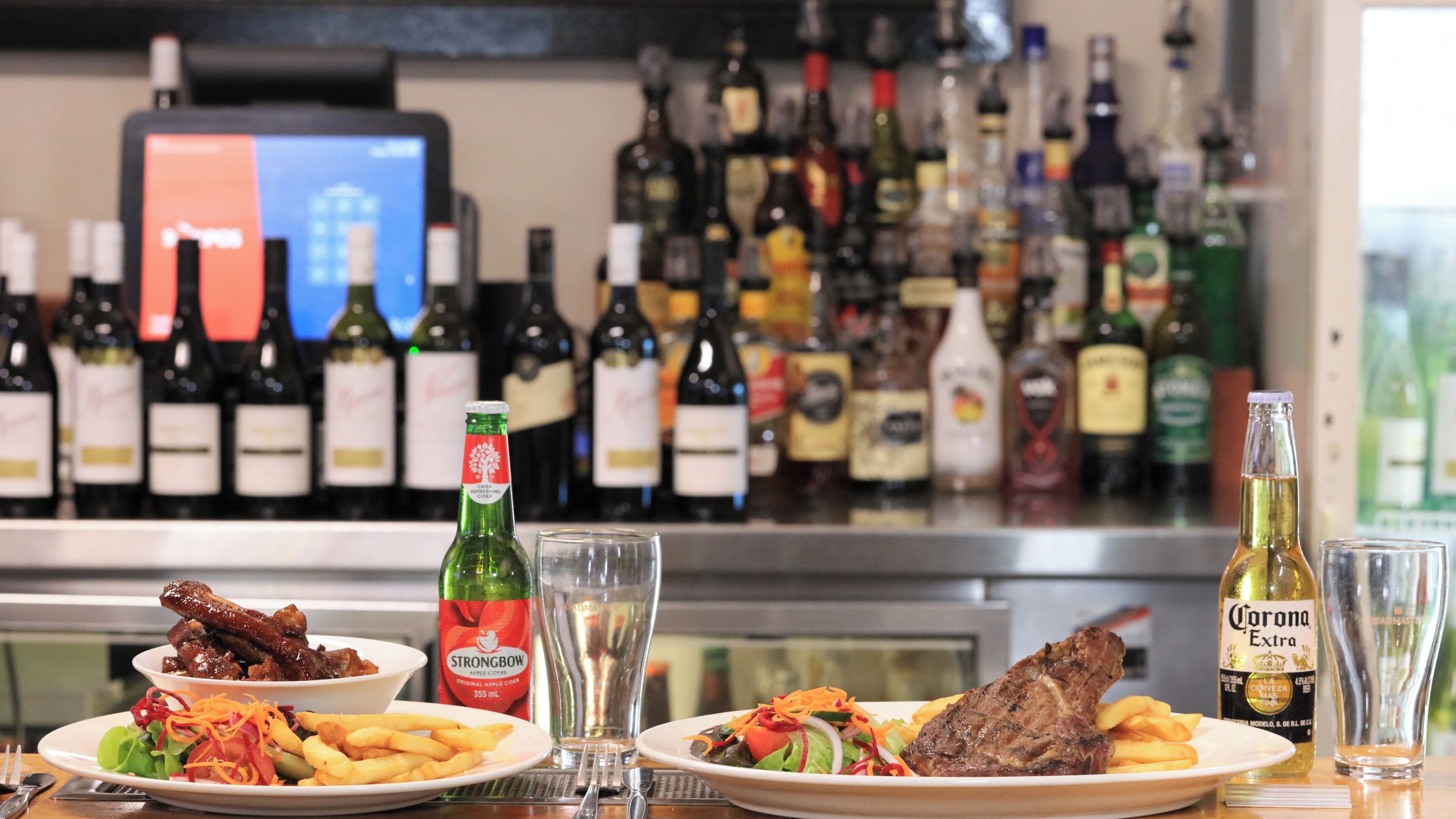 Uma área de bar com diversas garrafas de bebidas alcoólicas, uma garrafa de cidra Strongbow e pratos de comida, incluindo costelas e batatas fritas.