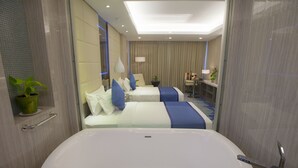 A modern hotel room with a large bathtub, two beds with blue and white bedding, a small desk, and a potted plant. - Grand Palace Hotel (Yangon)