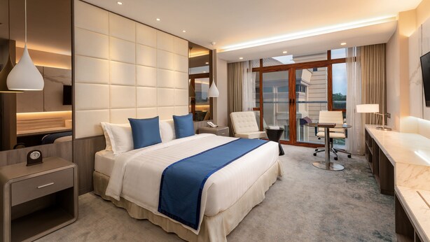A hotel room with a large bed, a desk, a chair, a TV, and a view of the city. - Grand Palace Hotel (Yangon)