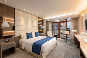 A hotel room with a large bed, a desk, a chair, a TV, and a view of the city. - Grand Palace Hotel (Yangon)