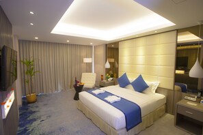 A modern hotel room with a large bed, a bedside table, a chair, a desk, a potted plant, and a TV. - Grand Palace Hotel (Yangon)