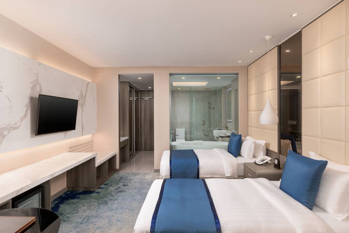 Habitación de hotel moderna con dos camas, televisor, escritorio y baño visible a través de una puerta de cristal.