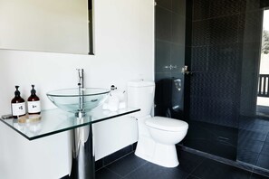 A modern bathroom with a glass sink, white toilet, and dark tiled walls. - Polperro Villas (Red Hill South)