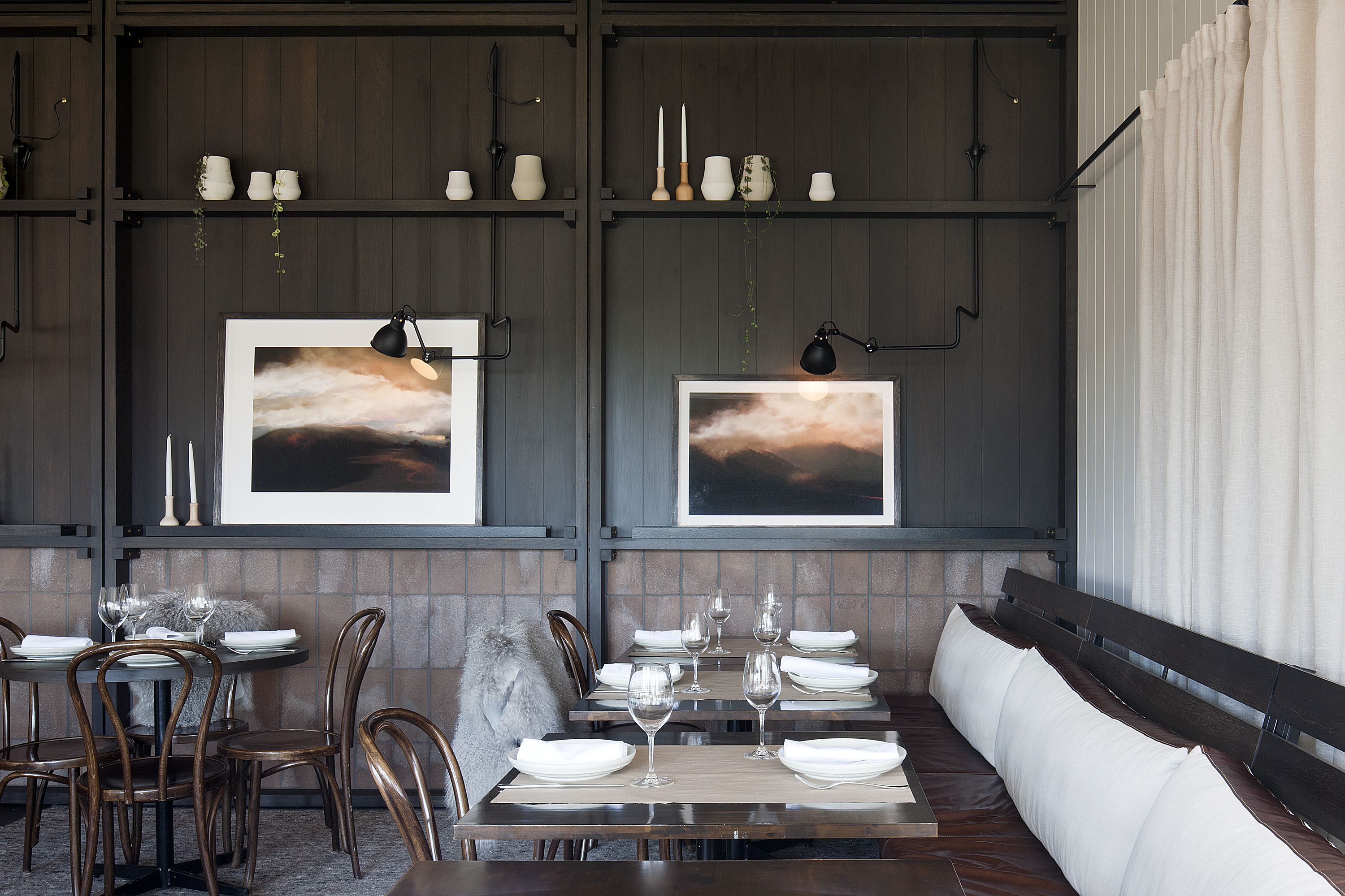 A dining area with a long wooden bench, a table set with white plates and glasses, and two framed abstract paintings on the wall.