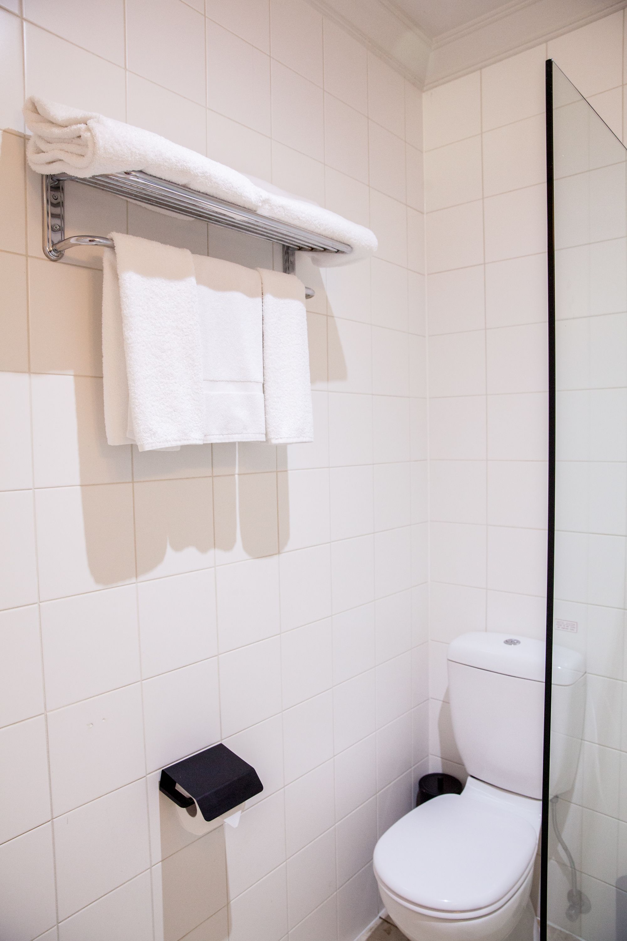 Un baño con azulejos blancos, un inodoro, un perchero con dos toallas y un dispensador de toallas de papel.
