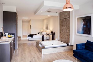 A modern hotel room with a kitchenette, a large bed, a sitting area with a blue sofa, and a bathroom with a bathtub. - Polperro Villas (Red Hill South)