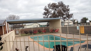 A fenced outdoor swimming pool with a covered area, surrounded by a metal fence and red chairs. - Highway Inn Motel (Hay)