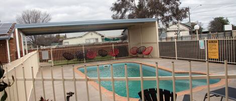 A fenced outdoor swimming pool with a covered area, surrounded by a metal fence and red chairs.