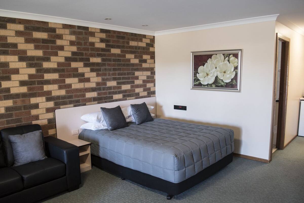 A bedroom with a bed, a nightstand, a chair, a brick-patterned wall, and a framed floral painting.