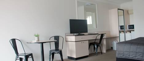 A hotel room with a bed, a desk with a computer and TV, two chairs, a small table with a plant, and a large mirror.