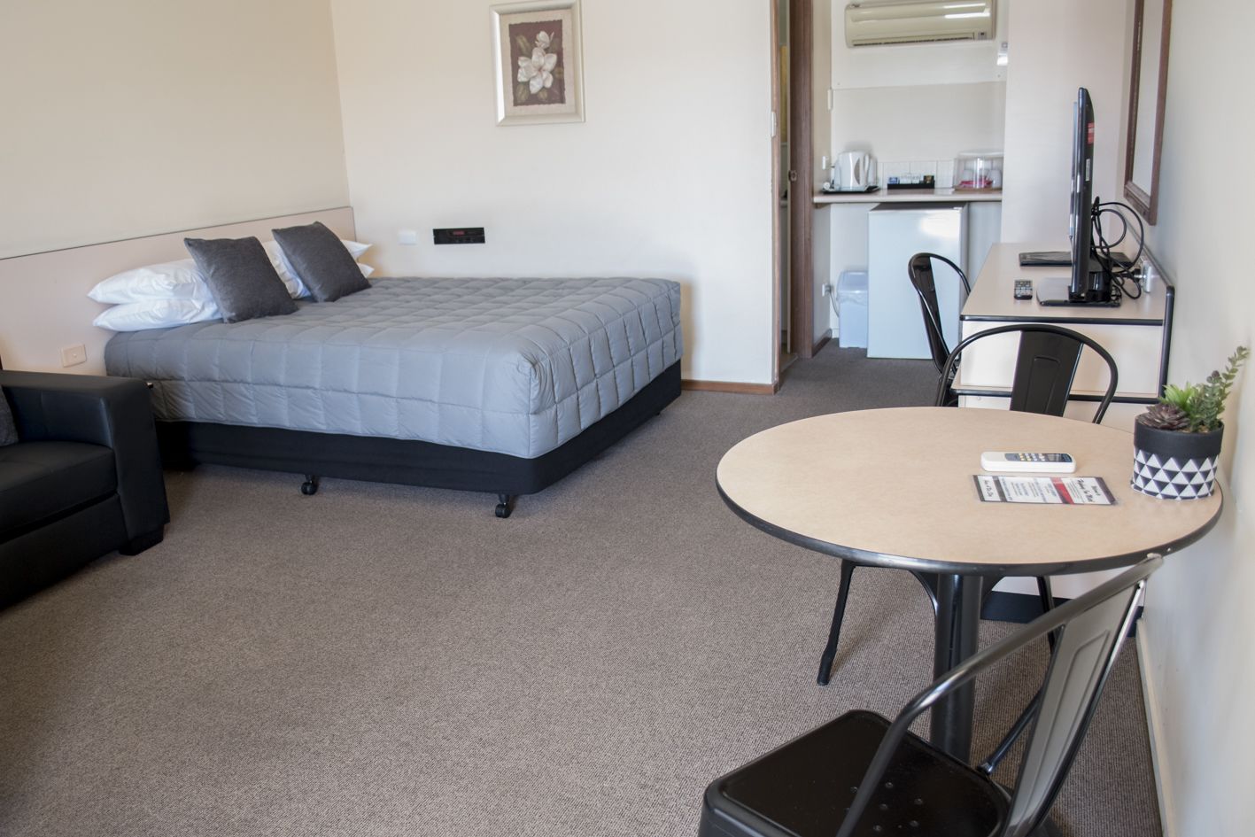 Habitación de hotel compacta con cama, un pequeño sofá, una mesa redonda con un control remoto y una planta en maceta.