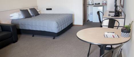 Habitación de hotel compacta con cama, un pequeño sofá, una mesa redonda con un control remoto y una planta en maceta.