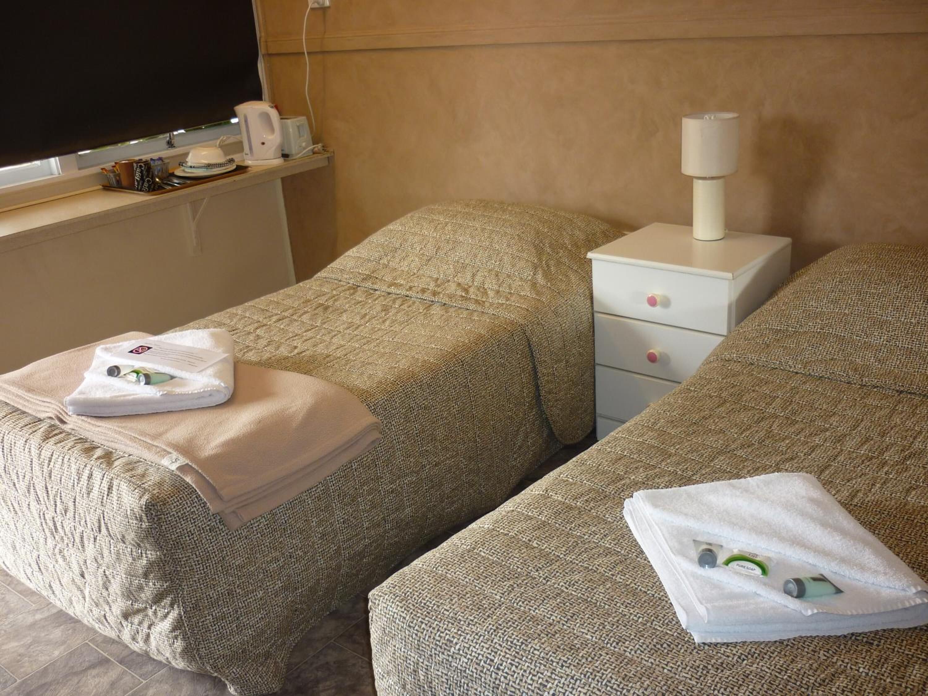 A hotel room with two beds, each with a towel and a bottle of shampoo. There is a nightstand with a lamp and a TV on the wall.