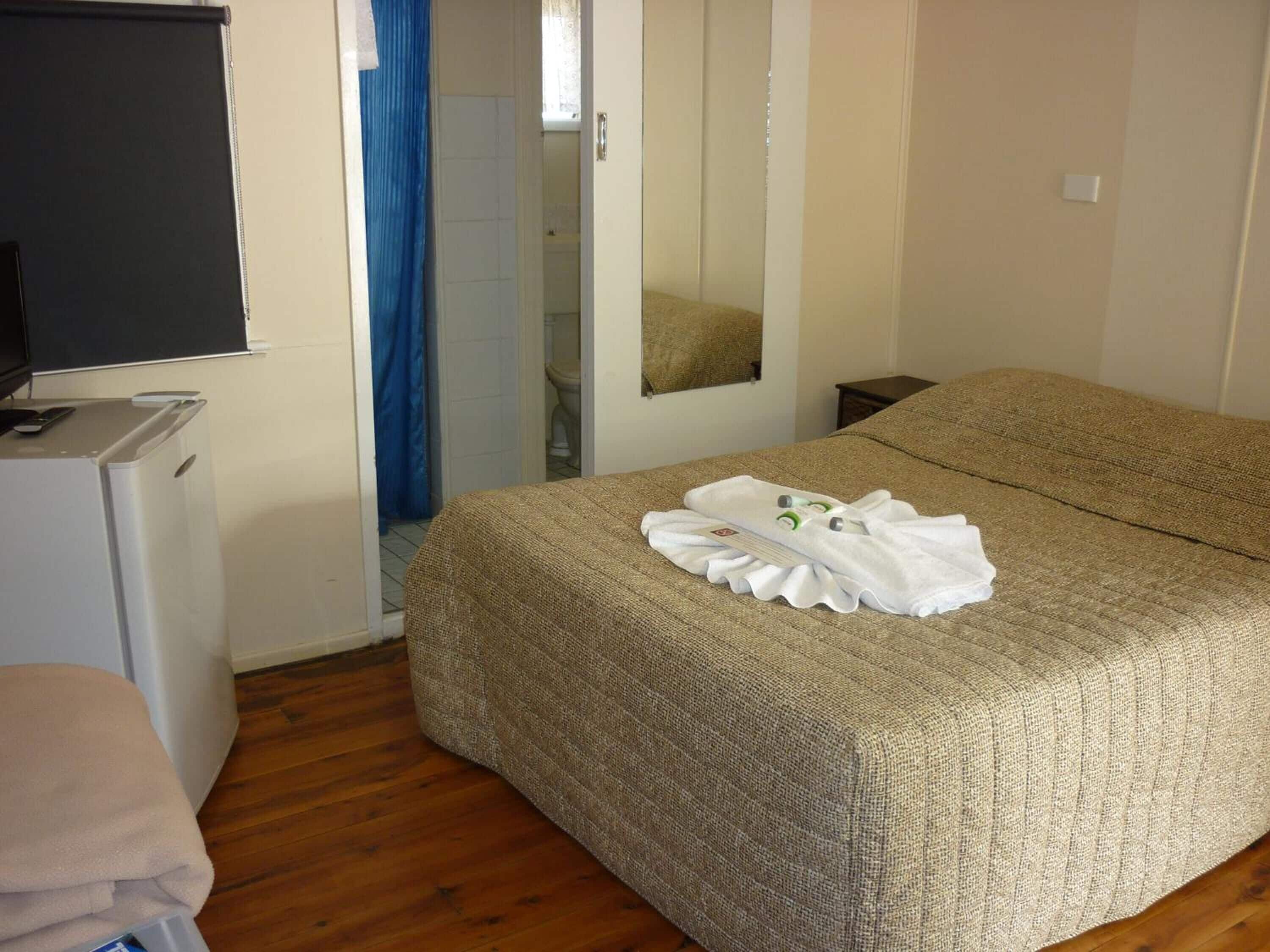 A hotel room with a bed, a small desk with a computer, a mirror, and a shower area.