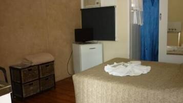 Una habitación pequeña y sencilla con una cama, un televisor sobre un mueble bajo, un tocador pequeño y una zona de ducha con cortina azul.