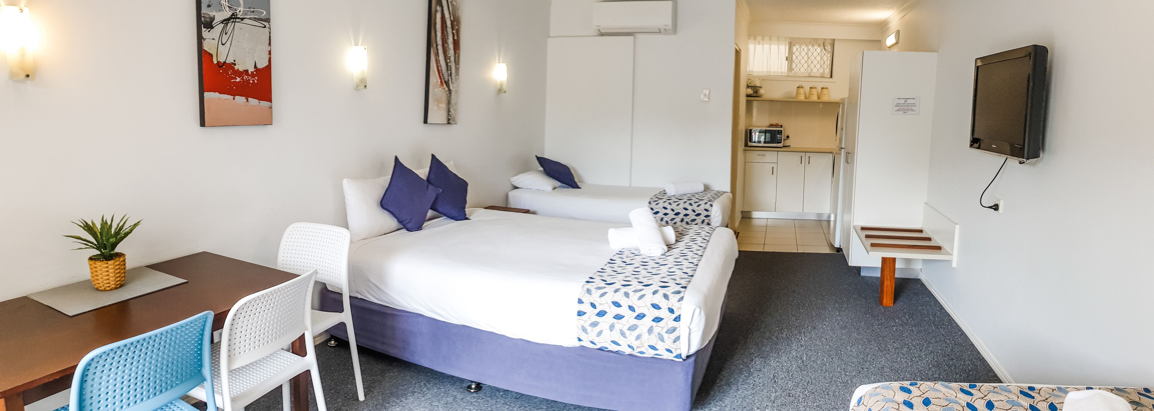 Une chambre d’hôtel comprenant un lit, un bureau, une chaise, une petite table avec une plante et un coin cuisine.