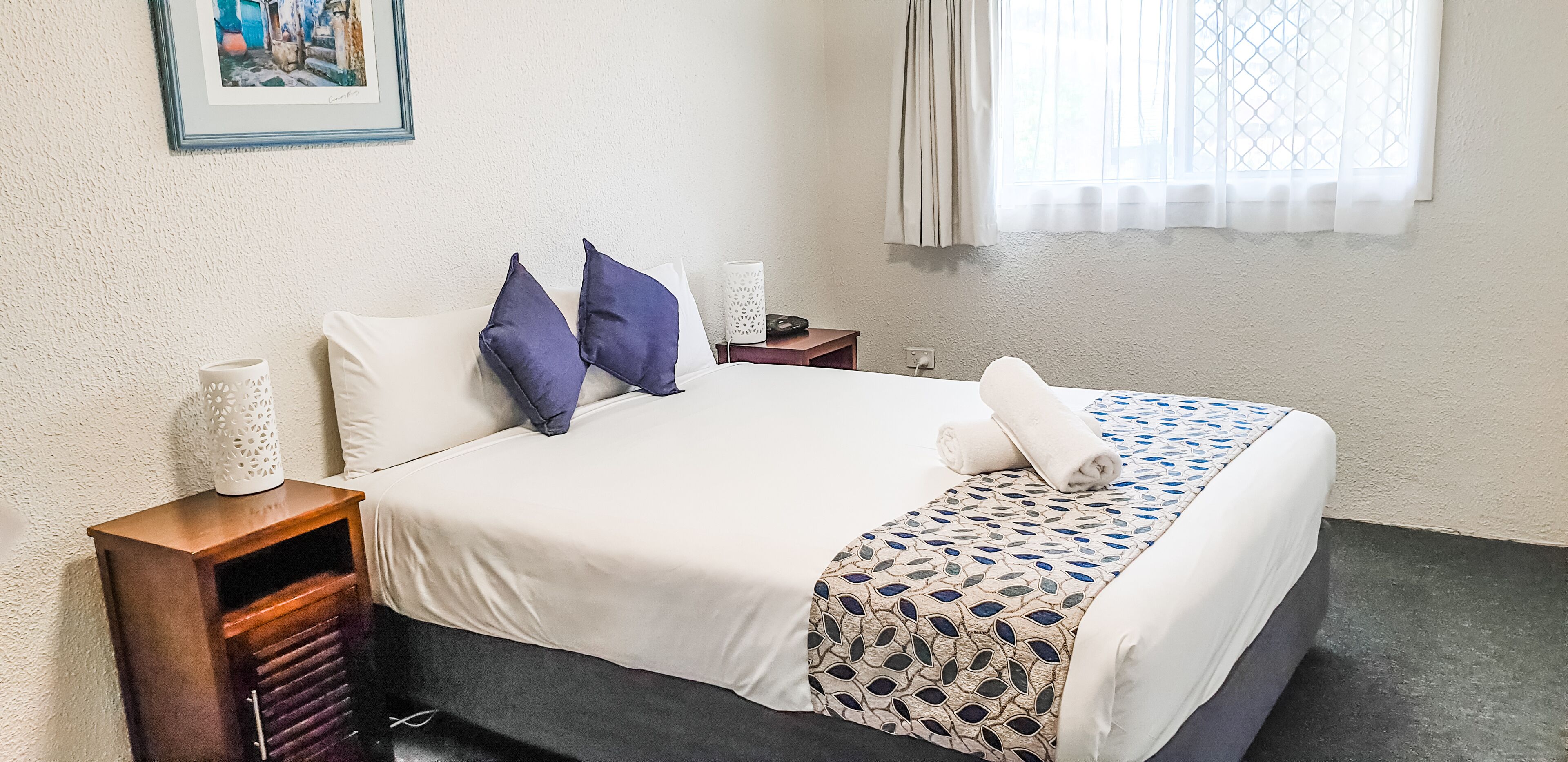 A neatly made bed with a patterned bedspread, two pillows, a bedside table with a lamp, and a framed picture on the wall.