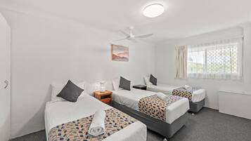Habitación de hotel con tres camas, ventilador de techo, un cuadro y una ventana con cortinas.