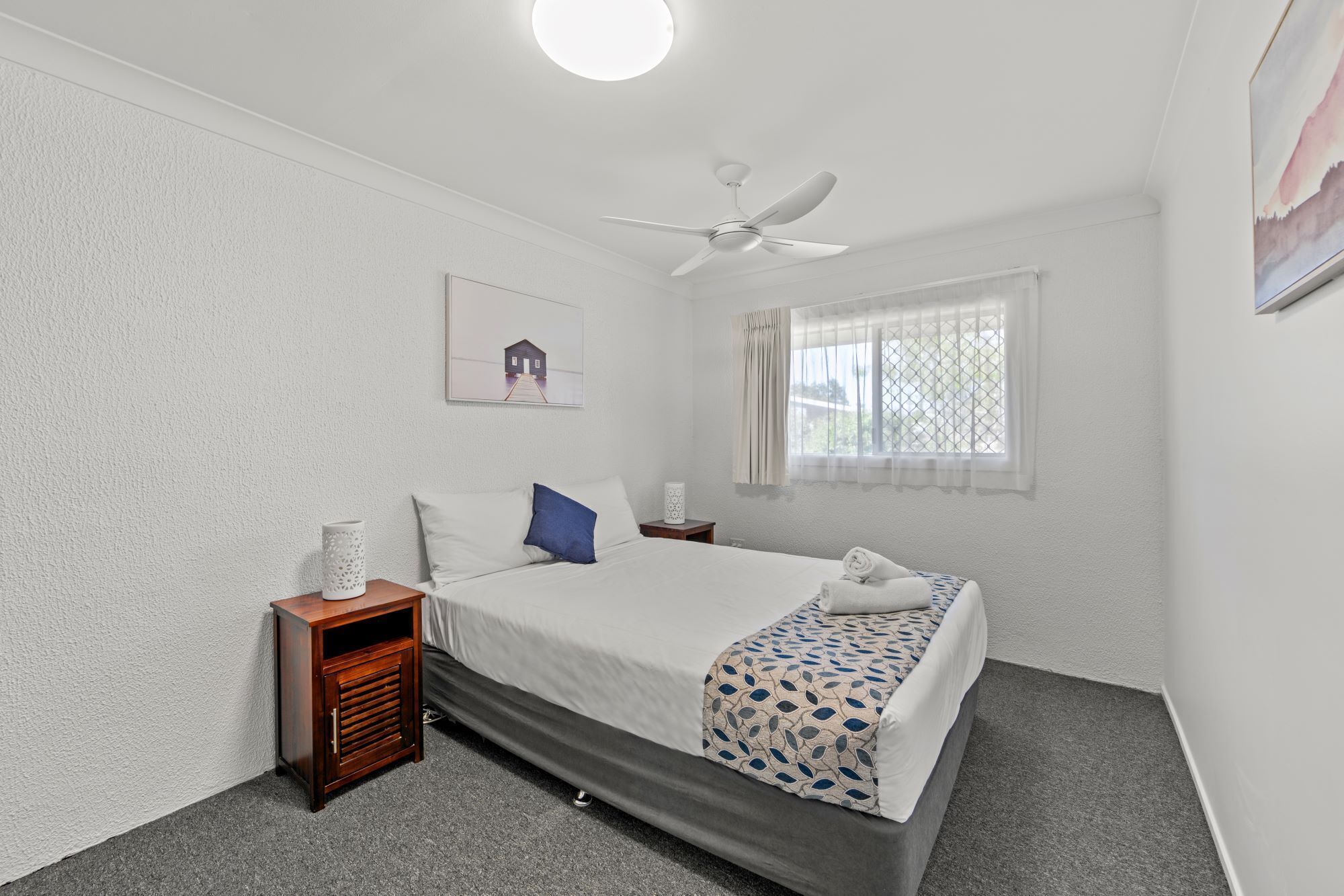 A bedroom with a bed, a nightstand, a ceiling fan, a window with curtains, and a wall-mounted picture.
