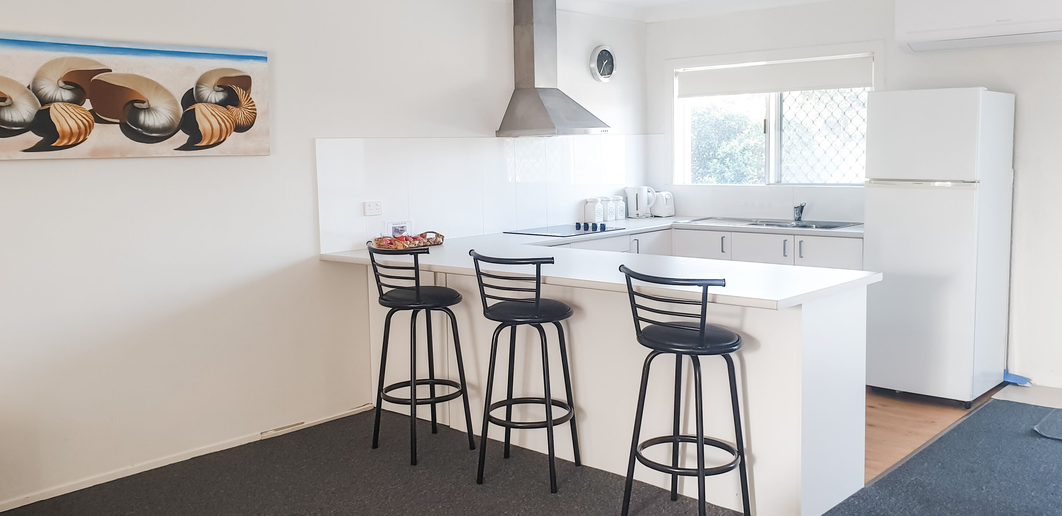 Una cocina moderna con encimera blanca, taburetes negros, una campana extractora de acero inoxidable y una ventana con vistas a zonas verdes.