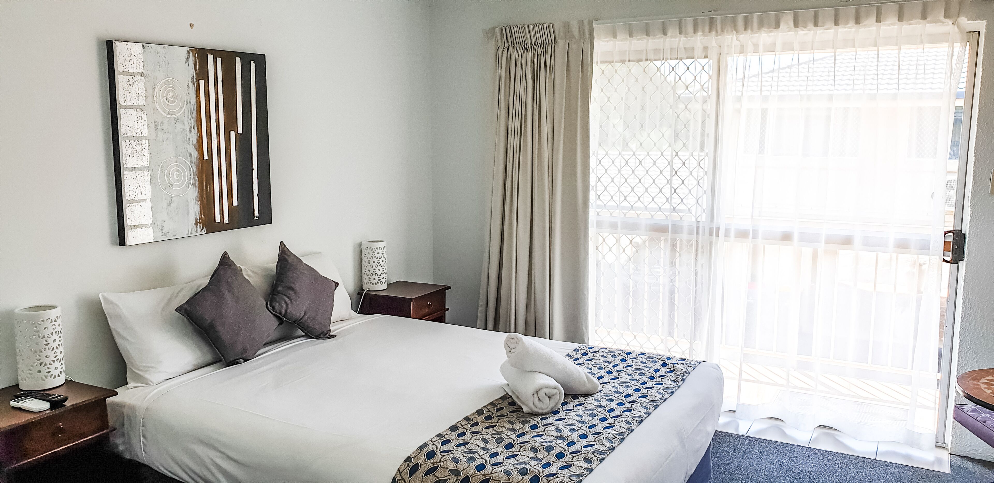 A neatly made bed with white and patterned bedding, a bedside table with a remote control, and a decorative wall piece.