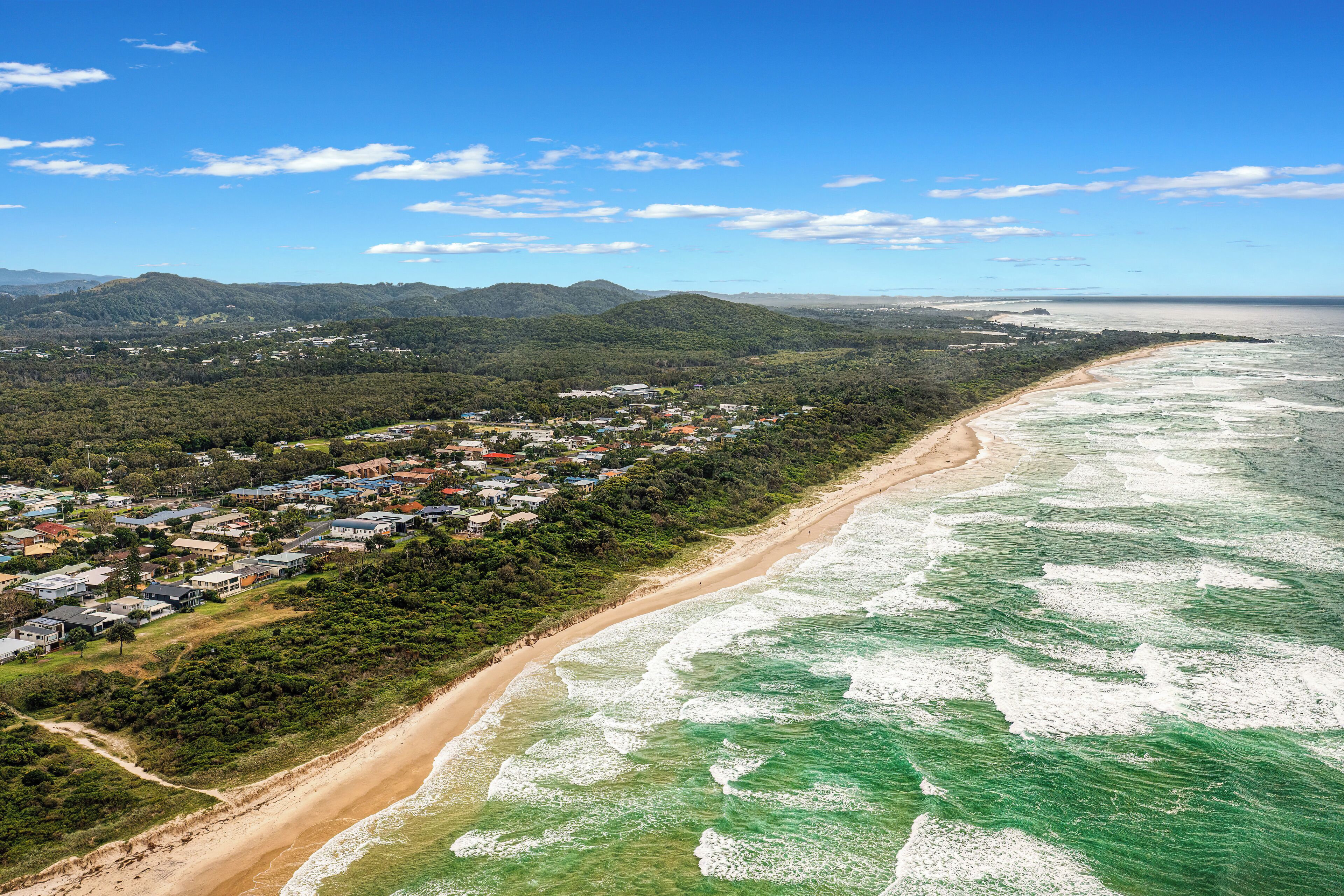 Vista aérea de un pueblo costero con playa, casas y colinas verdes al fondo.