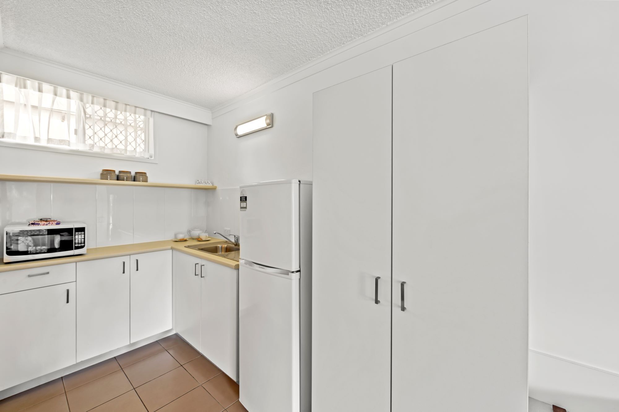 Eine Küche mit weißen Schränken, einer Mikrowelle, einem Kühlschrank und einem Fenster mit Vorhang.