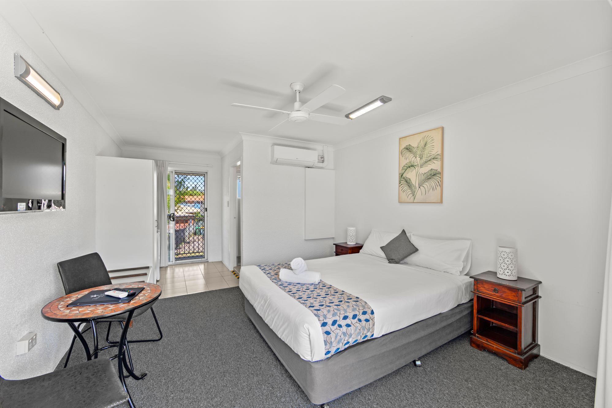 A hotel room with a bed, a desk, a chair, a ceiling fan, a painting, and an air conditioning unit.