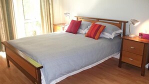 Una cama bien hecha con estructura de madera, una colcha gris y dos almohadas. Hay una mesita de noche de madera con una lámpara y una caja de pañuelos. El cuarto tiene piso de madera y una ventana con cortinas.