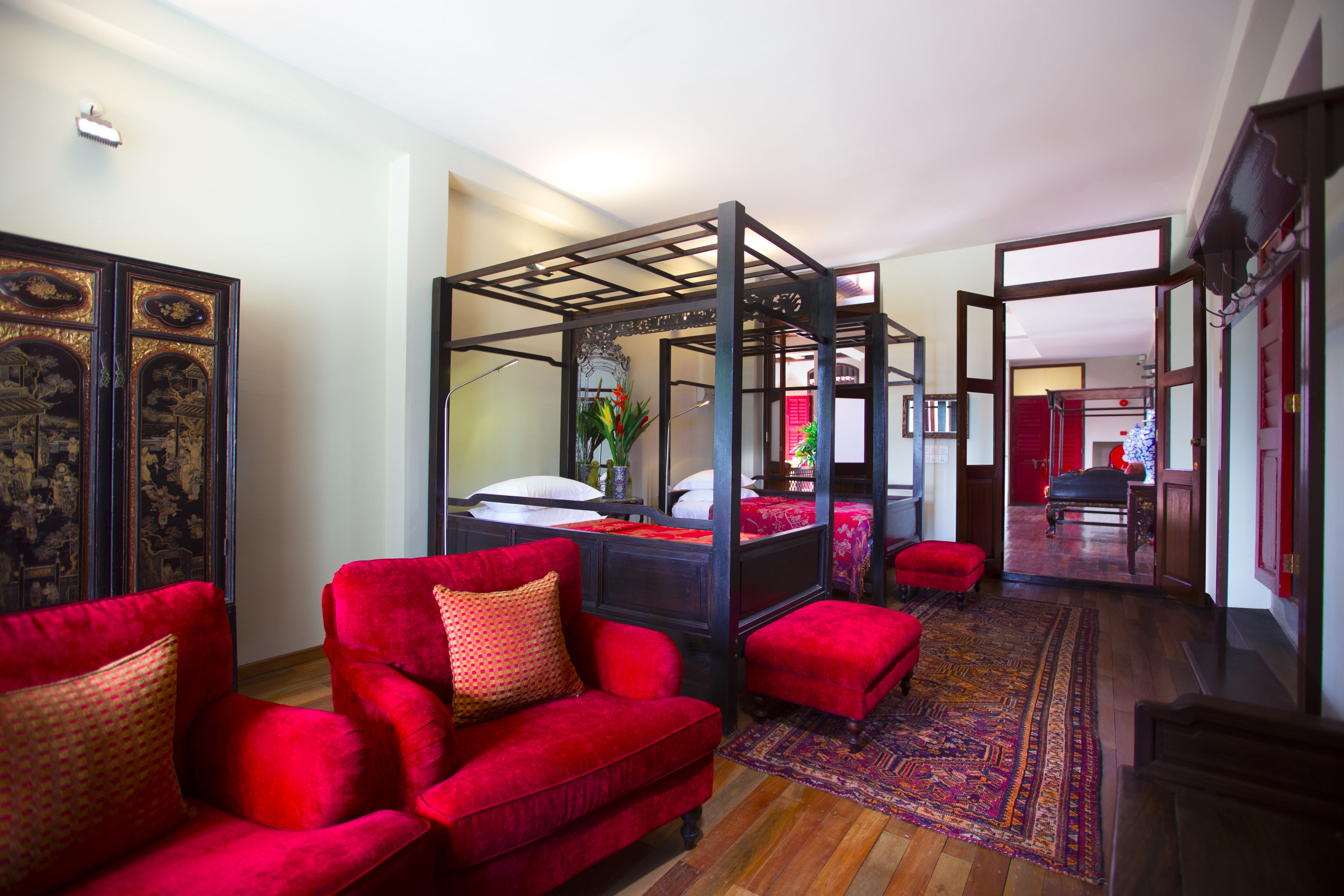 A traditional four-poster bed with a red upholstered headboard, a red sofa with patterned cushions, and a patterned rug on a wooden floor.