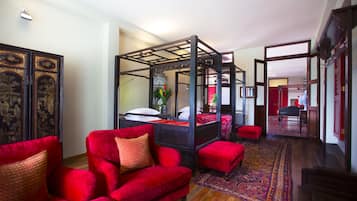 A traditional four-poster bed with a red upholstered headboard, a red sofa with patterned cushions, and a patterned rug on a wooden floor.