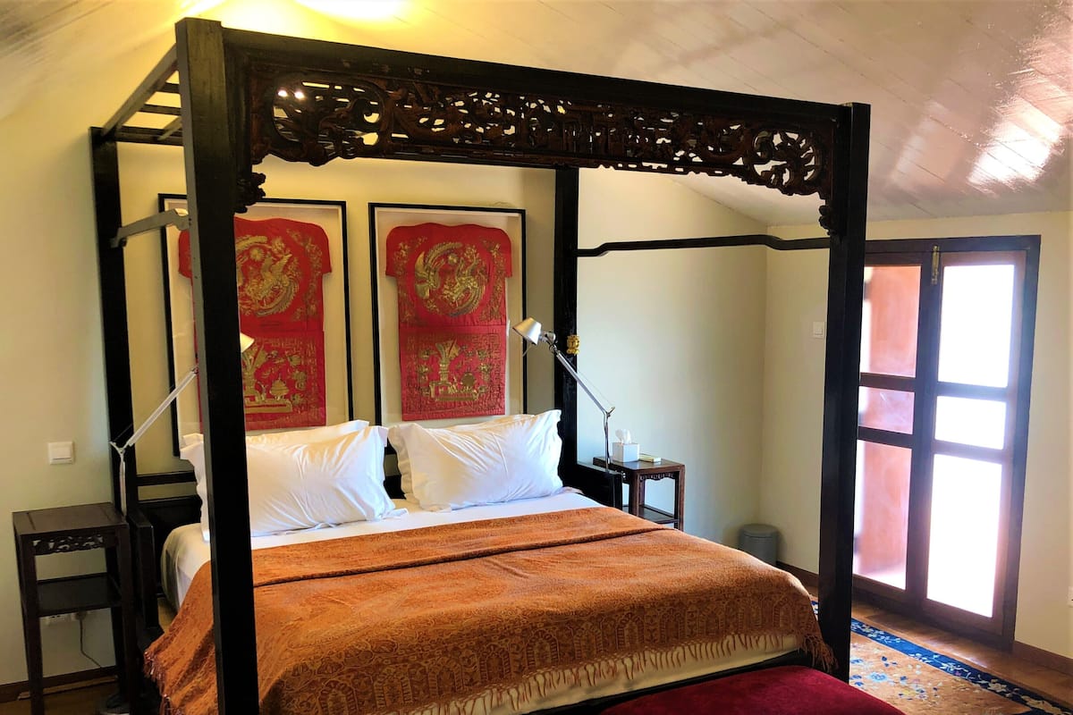 Un lit à baldaquin avec une couvre-lit rouge et blanche, une tête de lit en bois et un banc recouvert d’un tissu rouge.
