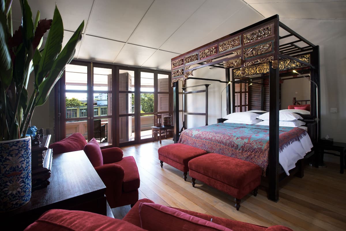 Un lit à baldaquin avec une couvre-lit à motifs, des fauteuils rembourrés de couleur rouge, une table en bois ornée d’un vase de fleurs et de grandes fenêtres donnant sur les bâtiments environnants.