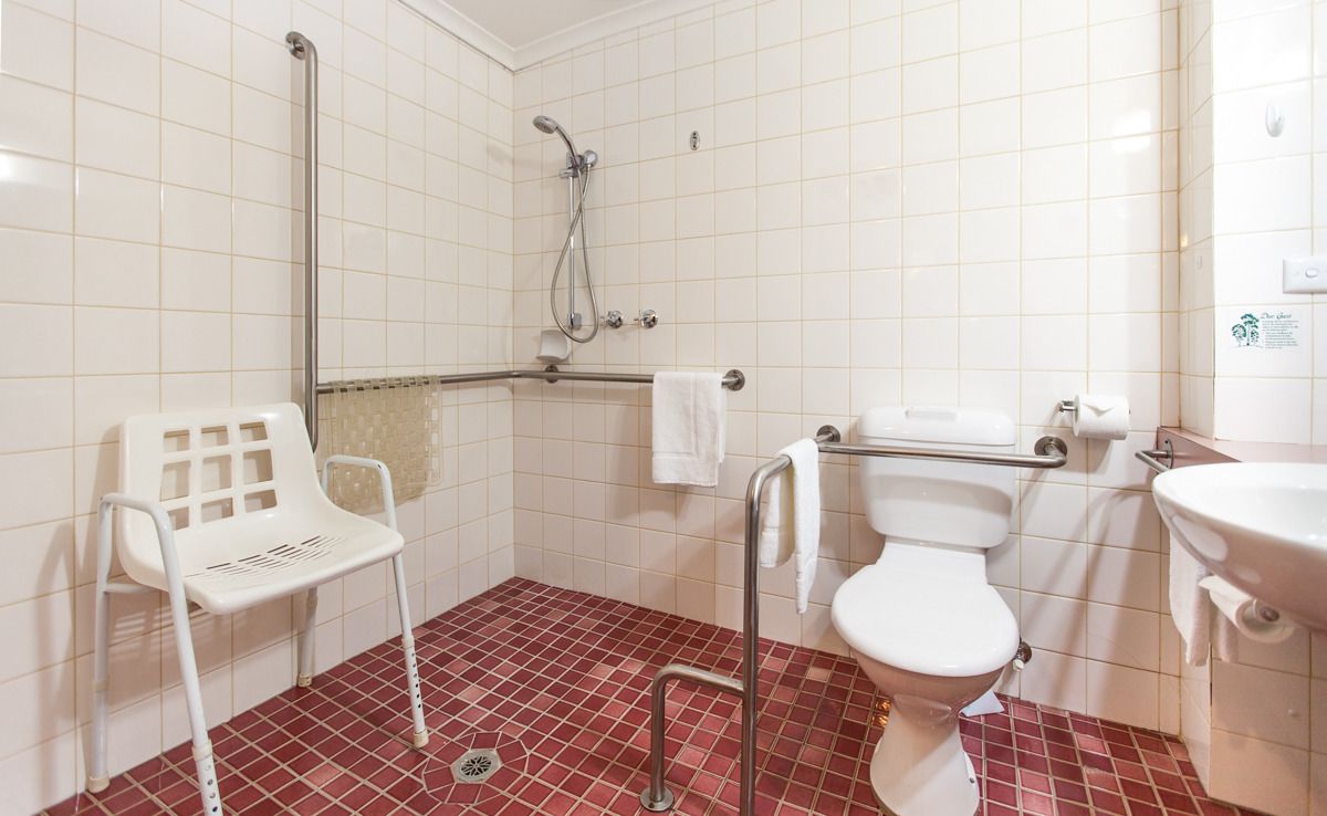 Un baño con inodoro blanco, ducha con cabezal de ducha manual, barra de apoyo, silla blanca y lavamanos.