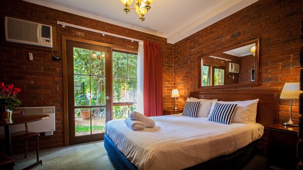 A hotel room with a brick wall, a large bed with white linens, a bedside table with a lamp, and a window with a view of greenery.