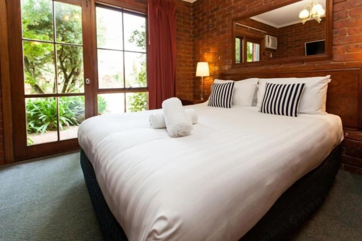 A hotel room with a large bed, a window with a view of greenery, and a brick wall.