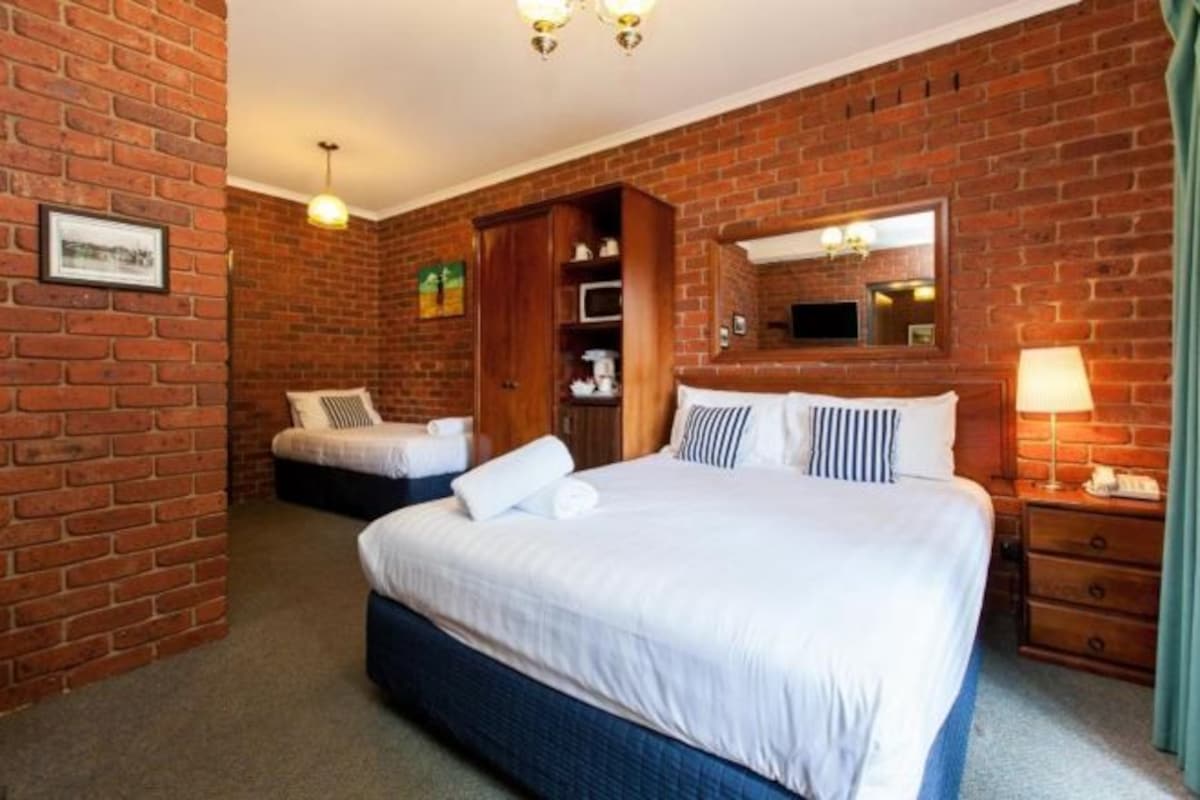 A hotel room with a brick wall, a large bed with white bedding, a bedside table with a lamp, and a wardrobe.