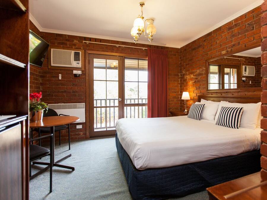 A hotel room with a brick wall, a large bed with white bedding, a desk with a chair, and a small table with a plant.