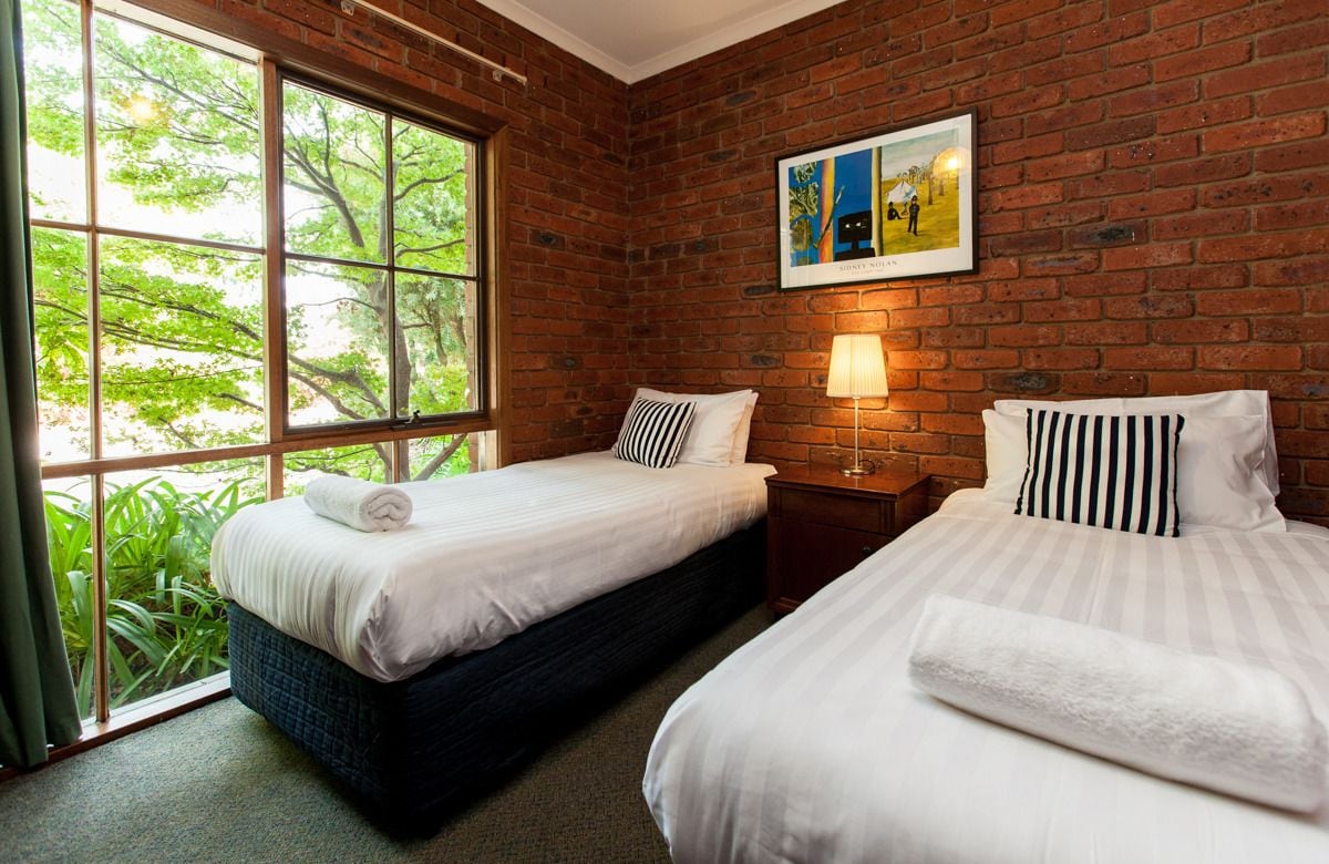 Une chambre d’hôtel avec deux lits, une grande fenêtre donnant sur un paysage verdoyant, un mur en briques et un tableau au-dessus des lits.