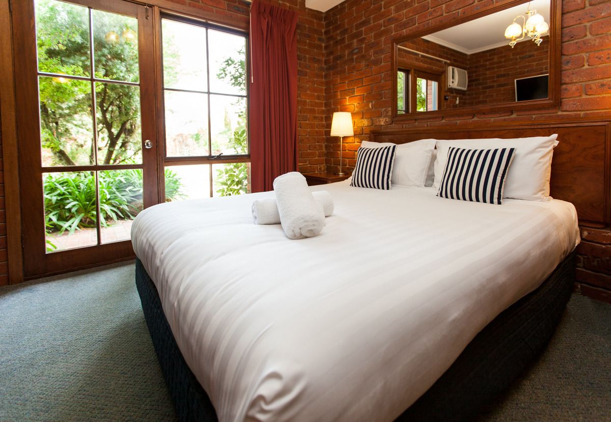 A hotel room with a large bed, a window with a view of greenery, a brick wall, and a mirror above the bed.