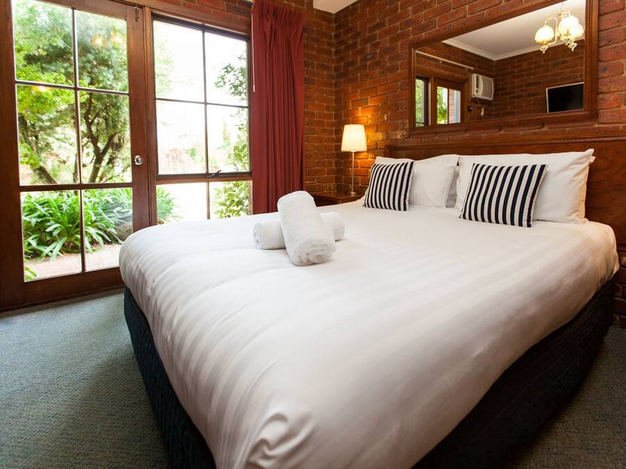 Une chambre d’hôtel avec un grand lit, une fenêtre donnant sur un paysage verdoyant, un mur en briques et un miroir au-dessus du lit.