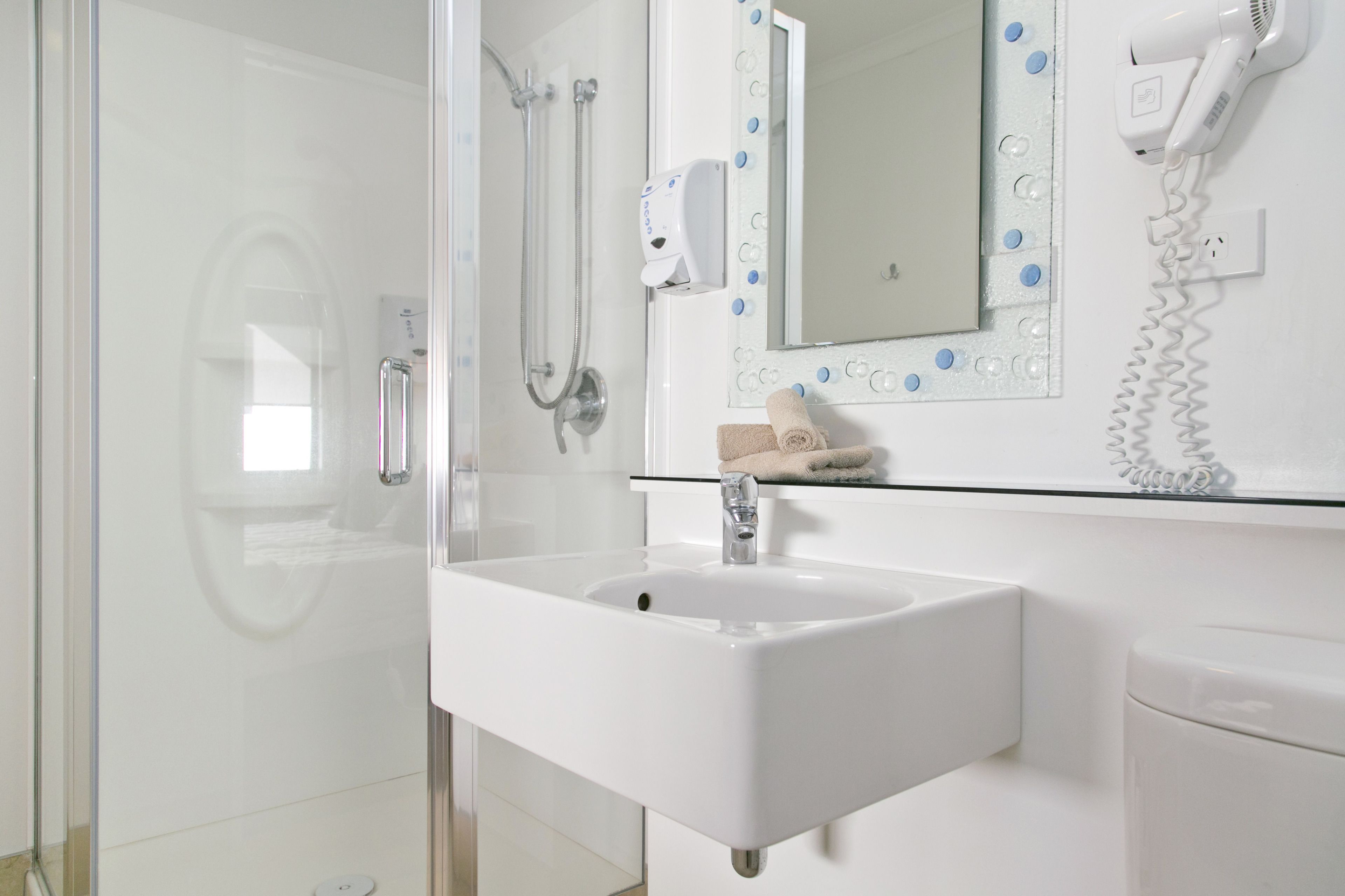 A bathroom with a glass shower enclosure, a white sink, a mirror with decorative blue dots, and a hairdryer on the wall.
