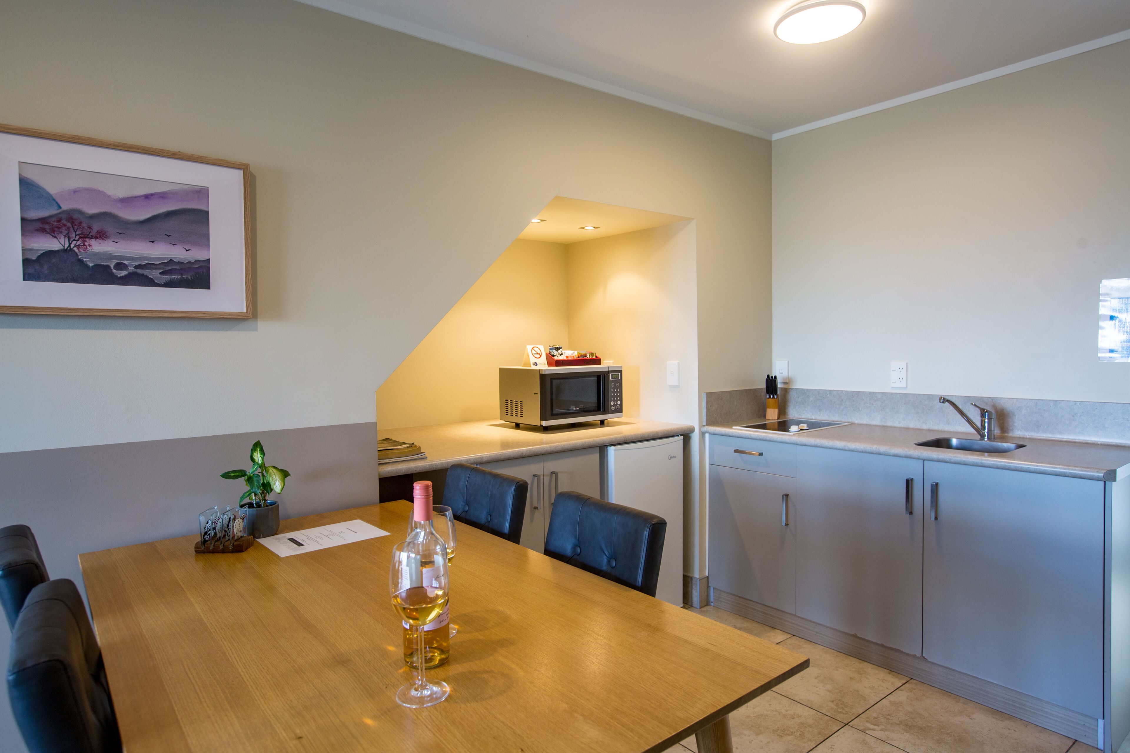 A compact kitchen with a microwave, sink, and cabinets. There is a dining table with chairs and a bottle of wine. A framed picture hangs on the wall.