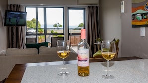 Une pièce avec un téléviseur accroché au mur, une table avec deux verres de vin et une bouteille, un canapé et une vue sur l’extérieur grâce à de grandes fenêtres.
