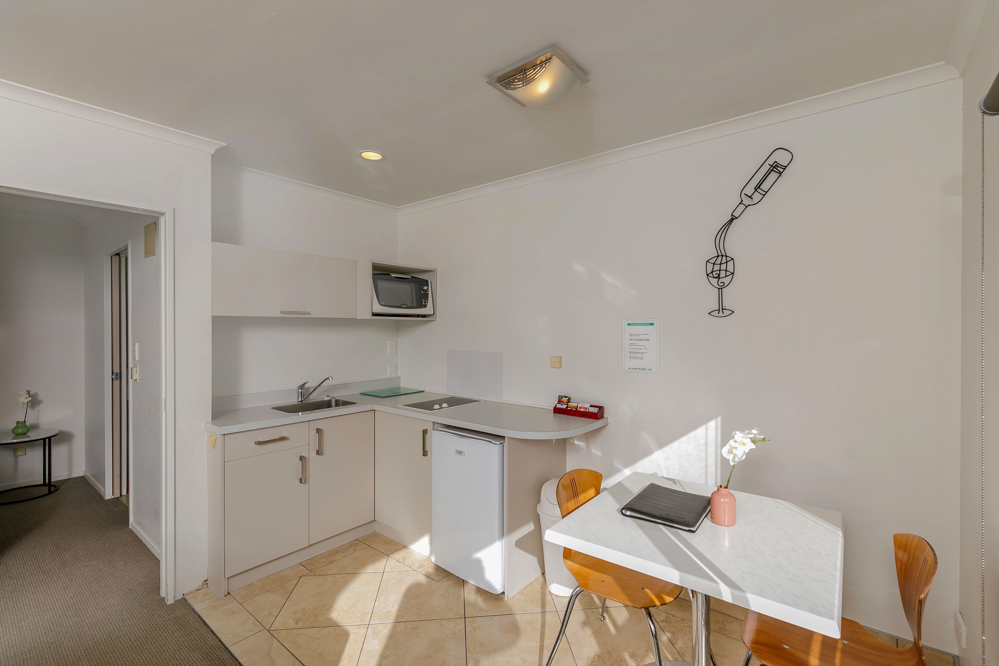 Uma cozinha compacta com micro-ondas, pia e um pequeno frigorífico. Há uma área de refeições com mesa e cadeiras, e um candeeiro fixado na parede.