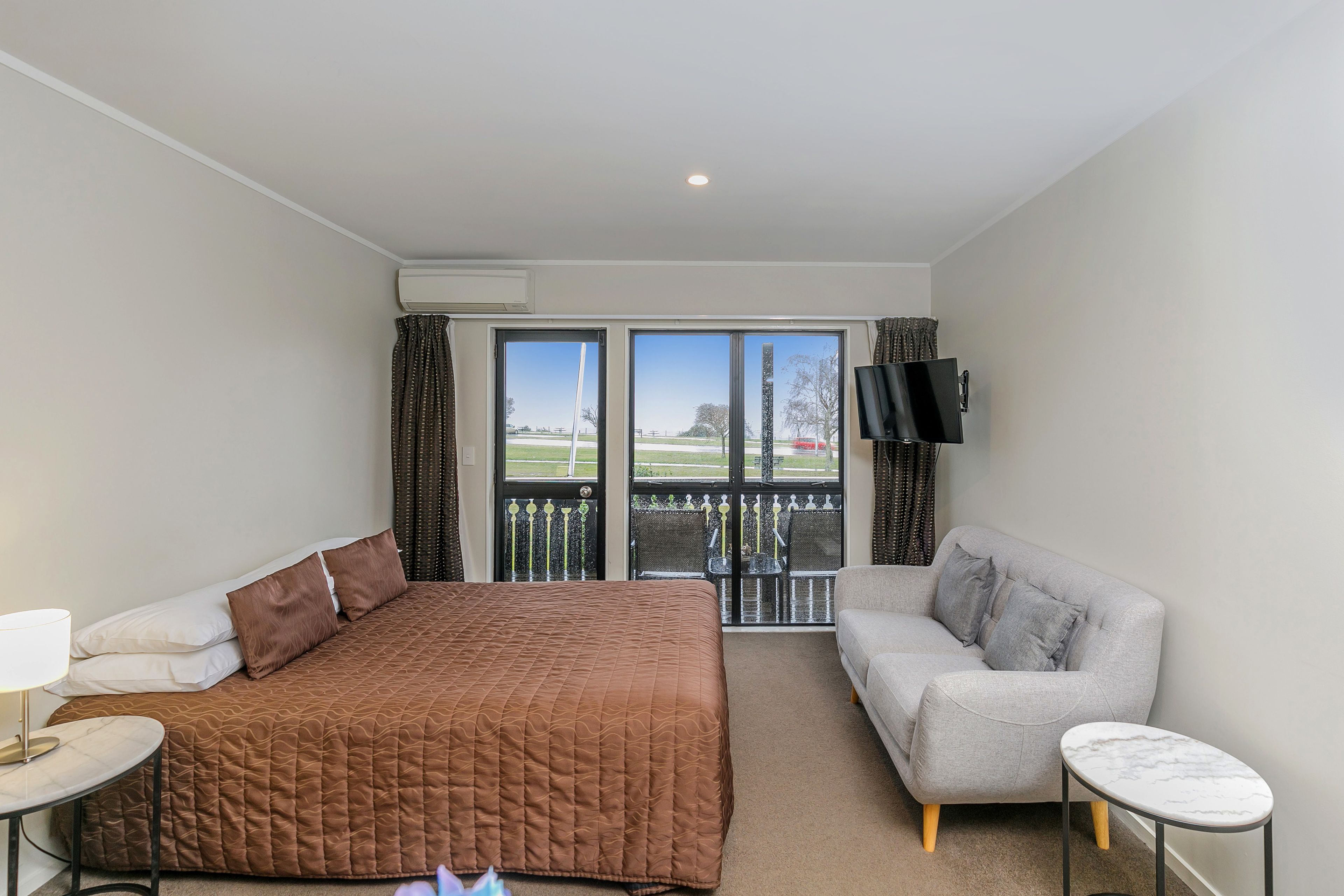 Une chambre d’hôtel avec un lit, un canapé gris, une petite table, une télévision et un balcon donnant sur l’extérieur.