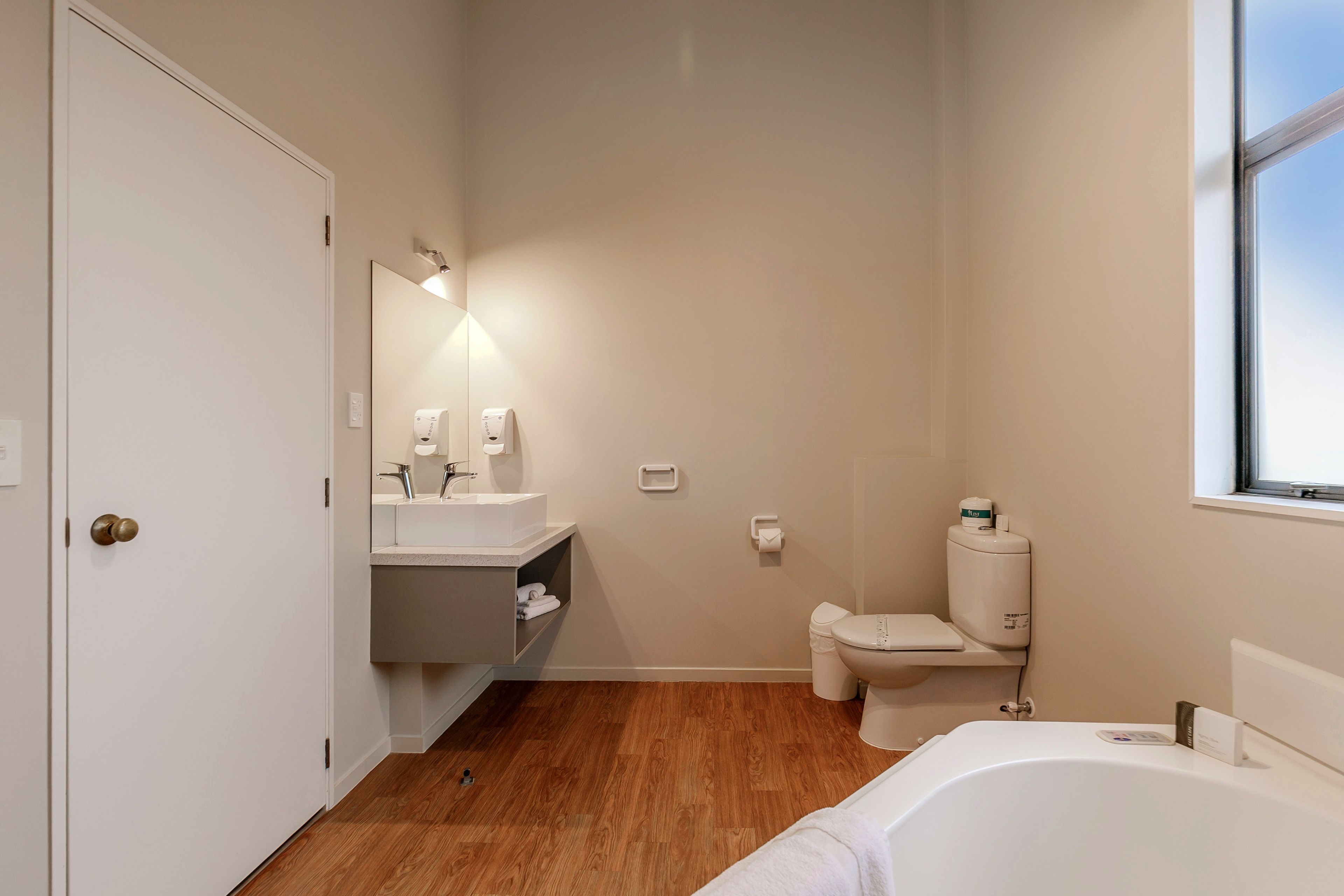 A modern bathroom with a wooden floor, a white bathtub, a toilet, and a sink with a mirror.